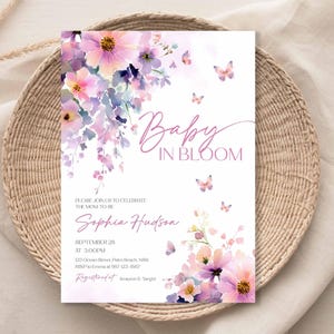 Może przedstawiać: Zaproszenie na baby shower z kwiatowym wzorem w odcieniach r&oacute;żu i fioletu, z napisem "Baby in Bloom". Zaproszenie zawiera szczeg&oacute;ły wydarzenia, takie jak data, godzina i miejsce.