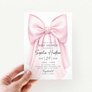 Op de afbeelding: Een uitnodiging voor een babyshower met een roze strik. De uitnodiging bevat de tekst "Doe mee voor een babyshower ter ere van Sophia Hudson" en evenementdetails. De uitnodiging wordt door een hand vastgehouden.