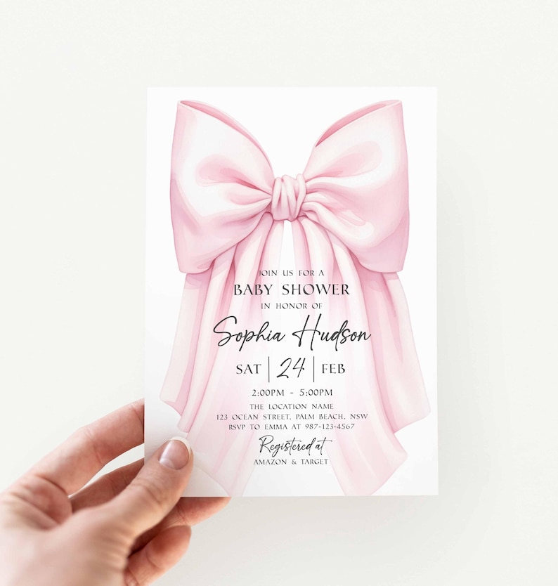 Op de afbeelding: Een uitnodiging voor een babyshower met een roze strik. De uitnodiging bevat de tekst "Doe mee voor een babyshower ter ere van Sophia Hudson" en evenementdetails. De uitnodiging wordt door een hand vastgehouden.