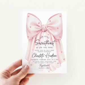 May include: A baby shower invitation featuring a large pink bow adorned with small pink hearts. The invitation reads "Our Little Sweetheart is on the way!" and includes details for the event honoring Charlotte Hudson.