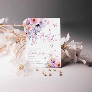 Może przedstawiać: Zaproszenie na baby shower z kwiatowym wzorem akwarelowym w odcieniach r&oacute;żu i fioletu. Tekst "Baby in Bloom" jest wyświetlany elegancką czcionką, wraz ze szczeg&oacute;łami wydarzenia. Elementy dekoracyjne obejmują motyle i małe koraliki.