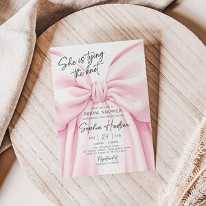K&ouml;nnte beinhalten: Eine Einladung zur Brautparty mit einem rosa Schleifendesign und dem Text "She is tying the knot". Die Einladung enth&auml;lt den Namen "Sophia Hudson" und Veranstaltungsdetails. Die Einladung liegt auf einer Holzoberfl&auml;che.