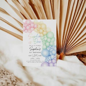 May include: A white invitation card with a colorful balloon arch design. The text reads "Sophia is One" and announces a first birthday celebration. The card includes the date, time, and location details.