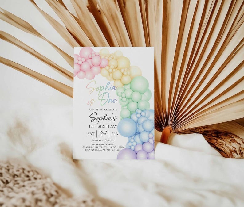 May include: A white invitation card with a colorful balloon arch design. The text reads "Sophia is One" and announces a first birthday celebration. The card includes the date, time, and location details.