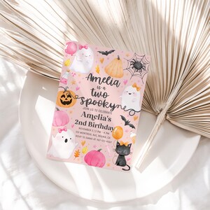 May include: A pink Halloween-themed invitation with the text "Amelia is a two spooky" and "Amelia's 2nd Birthday". The invitation features illustrations of ghosts, pumpkins, bats, and a black cat. The invitation is on a white plate.