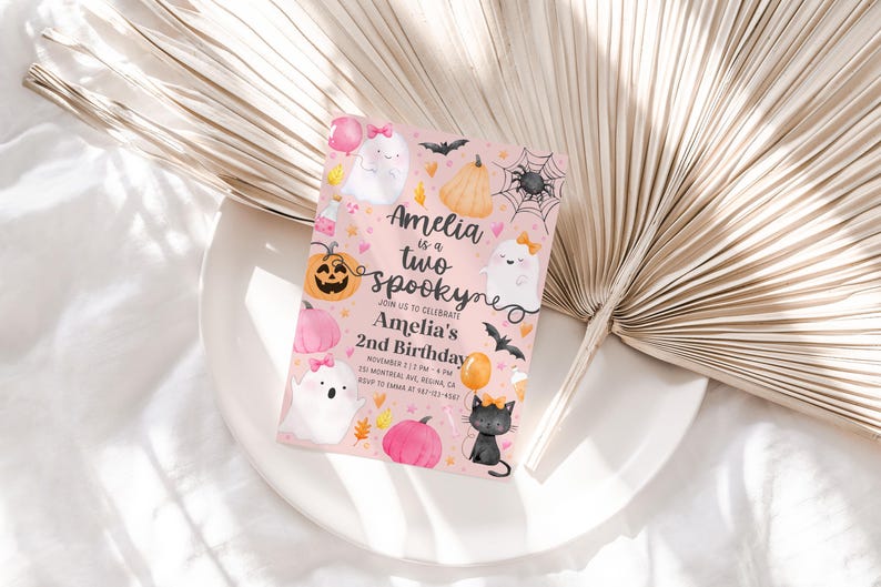 May include: A pink Halloween-themed invitation with the text "Amelia is a two spooky" and "Amelia's 2nd Birthday". The invitation features illustrations of ghosts, pumpkins, bats, and a black cat. The invitation is on a white plate.