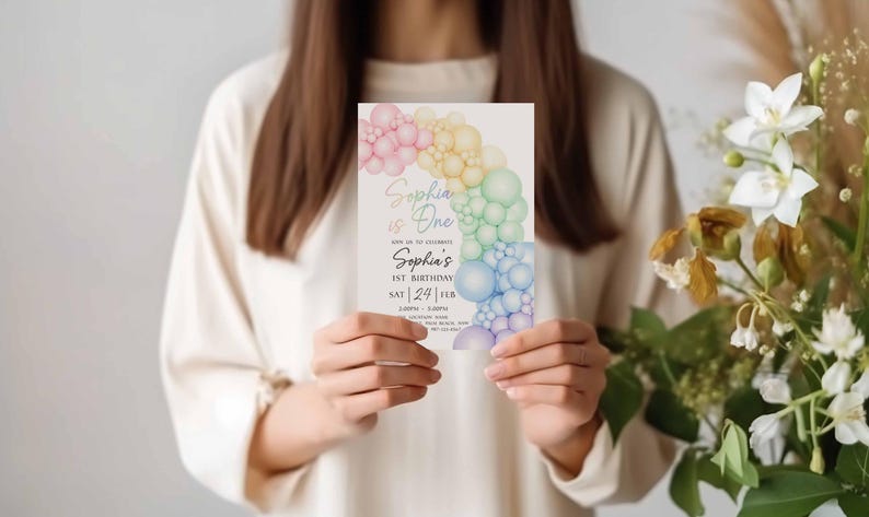 May include: A birthday invitation held by a person. The invitation features a rainbow-colored balloon arch design with the text "Sophia is One" and details for the celebration. The invitation is held in front of a neutral background.