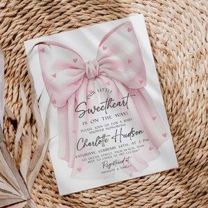 May include: Baby shower invitation with a watercolor bow design in shades of pink. The invitation includes the text "Our Little Sweetheart is on the way!" and details for the event, including the name Charlotte Hudson.