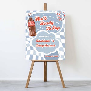 May include: A baby shower welcome sign on a wooden easel. The sign features a vintage-style design with a brown soda bottle and the text "She's Ready To Pop" and "Welcome to Charlotte's Baby Shower". The background has a blue and white checkered pattern.