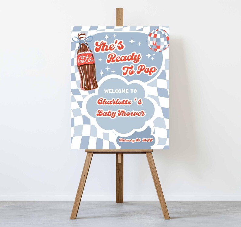 May include: A baby shower welcome sign on a wooden easel. The sign features a vintage-style design with a brown soda bottle and the text "She's Ready To Pop" and "Welcome to Charlotte's Baby Shower". The background has a blue and white checkered pattern.