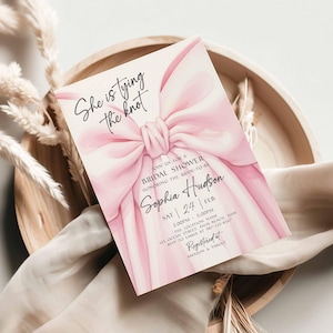 K&ouml;nnte beinhalten: Eine Einladung zur Brautparty mit einem gro&szlig;en rosa Schleifendesign und dem Text "She is tying the knot". Die Einladung enth&auml;lt Details f&uuml;r Sophia Hudsons Brautparty, einschlie&szlig;lich Datum, Uhrzeit und Ort. Die Einladung liegt auf einem Holztablett.