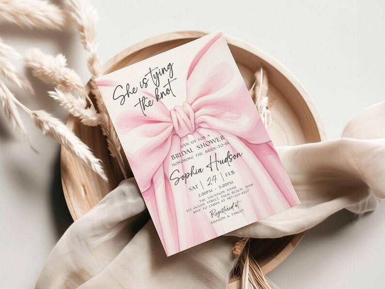 K&ouml;nnte beinhalten: Eine Einladung zur Brautparty mit einem gro&szlig;en rosa Schleifendesign und dem Text "She is tying the knot". Die Einladung enth&auml;lt Details f&uuml;r Sophia Hudsons Brautparty, einschlie&szlig;lich Datum, Uhrzeit und Ort. Die Einladung liegt auf einem Holztablett.