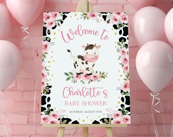 May include: Welcome to Charlotte's Baby Shower sign with a pink and black cow print background. The sign features a pink and white cow with a pink nose and black eyes. The text on the sign is in black and gold.