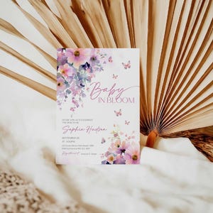 Może przedstawiać: Zaproszenie na baby shower z ilustracjami kwiat&oacute;w i motyli. Zaproszenie zawiera tekst "Baby in Bloom" oraz szczeg&oacute;ły wydarzenia, w tym imię Sophia Hudson oraz datę i godzinę. Projekt zawiera kolory r&oacute;żowy, fioletowy i biały.