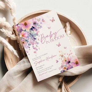 Może przedstawiać: Zaproszenie na baby shower z napisem "Baby in Bloom" w delikatnej czcionce. Projekt przedstawia akwarelowe kwiaty w odcieniach r&oacute;żu, fioletu i ż&oacute;łci, z małymi motywami motyli. Zaproszenie jest na kremowym tle.