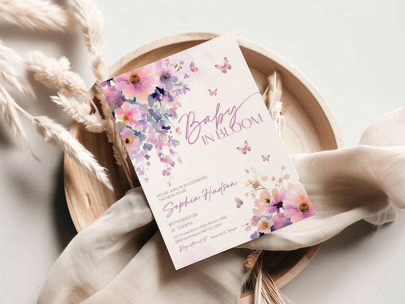 Może przedstawiać: Zaproszenie na baby shower z napisem "Baby in Bloom" w delikatnej czcionce. Projekt przedstawia akwarelowe kwiaty w odcieniach r&oacute;żu, fioletu i ż&oacute;łci, z małymi motywami motyli. Zaproszenie jest na kremowym tle.