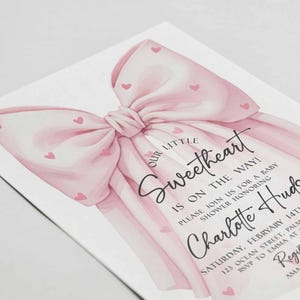 May include: A baby shower invitation featuring a large pink bow with small heart accents. The text reads "Our Little Sweetheart is on the way!" and includes details for the event, including the name "Charlotte Hudson".