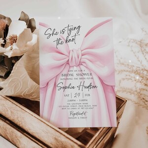 K&ouml;nnte beinhalten: Eine Einladung zur Brautparty mit einem gro&szlig;en rosa Schleifendesign und dem Text "She is tying the knot". Die Einladung enth&auml;lt den Namen der Braut, das Datum, die Uhrzeit und den Ort. Die Einladung befindet sich in einer Holzkiste mit Blumen.