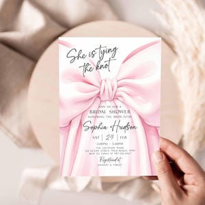 K&ouml;nnte beinhalten: Eine rosa Brautparty-Einladung mit dem Text "She is tying the knot" und Details f&uuml;r Sophias Dusche. Die Einladung zeigt ein gro&szlig;es rosa Schleifendesign und wird von einer Hand gehalten.