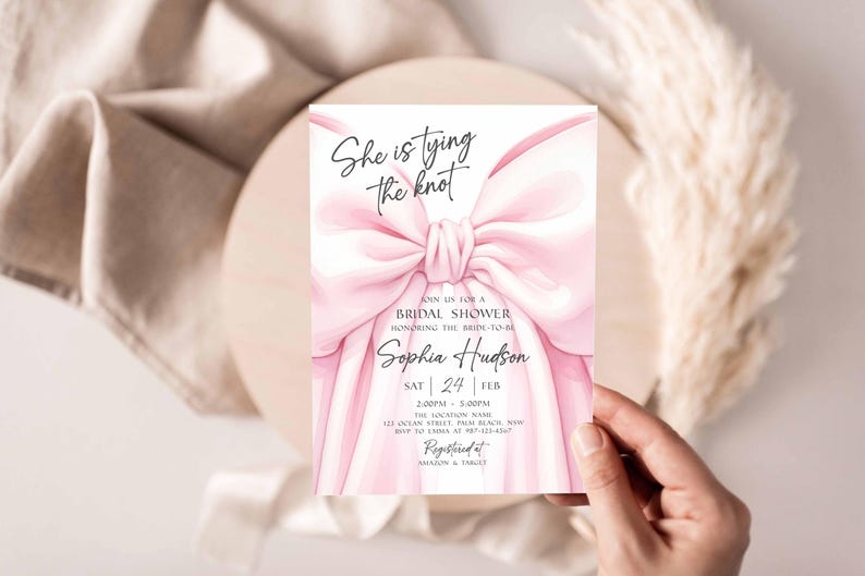 K&ouml;nnte beinhalten: Eine rosa Brautparty-Einladung mit dem Text "She is tying the knot" und Details f&uuml;r Sophias Dusche. Die Einladung zeigt ein gro&szlig;es rosa Schleifendesign und wird von einer Hand gehalten.
