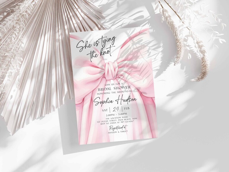 K&ouml;nnte beinhalten: Einladungskarte zur Brautparty mit rosa Schleife und dem Text "She is tying the knot". Die Einladung enth&auml;lt den Namen der Braut, Datum und Uhrzeit. Registriert bei Amazon & Target.