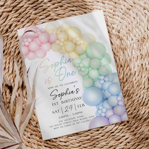 May include: A white invitation card with a pastel rainbow balloon arch design. The text "Sophia is One" is written in a script font, along with details for a first birthday celebration.