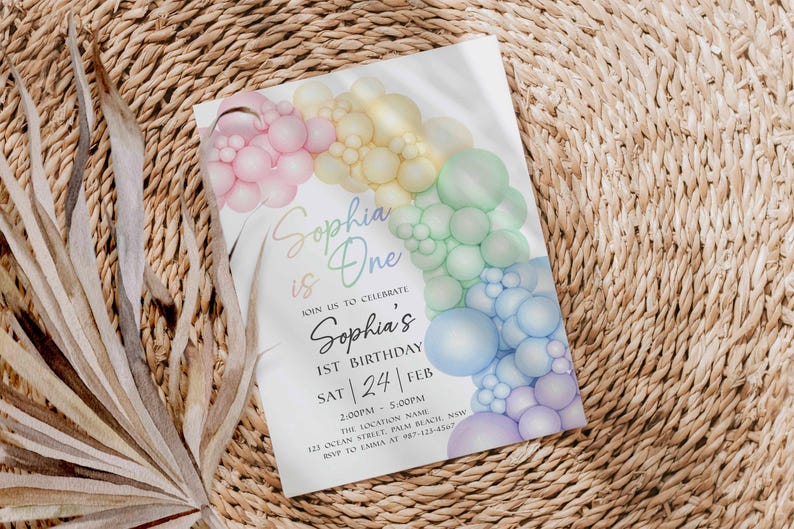 May include: A white invitation card with a pastel rainbow balloon arch design. The text "Sophia is One" is written in a script font, along with details for a first birthday celebration.
