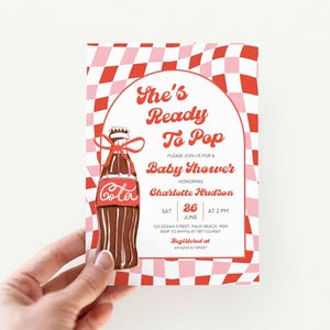 May include: Baby shower invitation with a red and pink checkered background. The invitation features a brown cola bottle illustration with a red ribbon. Text reads "She's Ready To Pop" and details for the event.