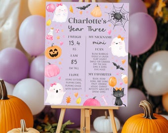 May include: A decorative sign on an easel with a purple background, featuring the text "Charlotte's Year Three." The sign includes illustrations of pumpkins, ghosts, and a black cat. The sign also includes the child's weight, height, nickname, and favorite things.