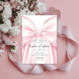 May include: A baby shower invitation with a large, soft pink bow design. The invitation includes the text "Join us for a Baby Shower in Honor of Sophia Hudson's" and event details. The background is a soft pink with flowers and ribbon.