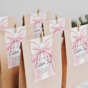 May include: Several kraft paper gift bags with custom tags. Each tag features a watercolor pink bow design and the words "Custom Tag". The tags are held in place with small wooden clothespins.