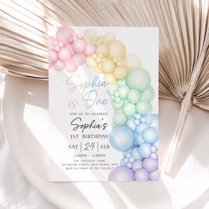 May include: A pastel rainbow-themed birthday invitation. The invitation features a colorful balloon arch and the text "Sophia is One." The date is Saturday, February 24th, with the time and location details below. The design is soft and whimsical.