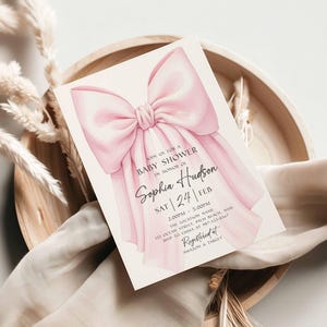 Op de afbeelding: Een uitnodiging voor een babyshower met een roze strik. De kaart bevat de tekst "Join us for a Baby Shower in Honor of Sophia Hudson" en evenementdetails. De uitnodiging ligt op een houten dienblad met decoratieve elementen.