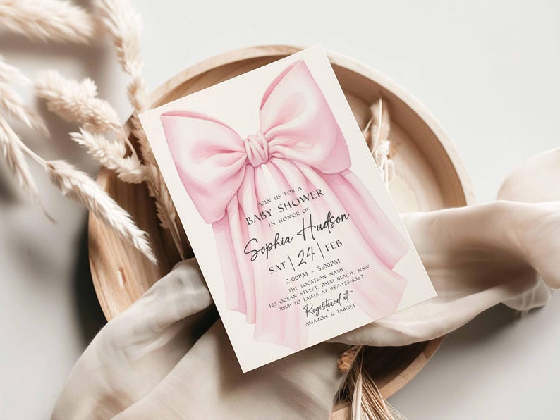 Op de afbeelding: Een uitnodiging voor een babyshower met een roze strik. De kaart bevat de tekst "Join us for a Baby Shower in Honor of Sophia Hudson" en evenementdetails. De uitnodiging ligt op een houten dienblad met decoratieve elementen.