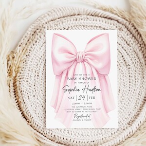 Op de afbeelding: Een uitnodiging voor een babyshower met een grote, zachte roze strik. De uitnodiging bevat de tekst "Doe mee voor een babyshower ter ere van Sophia Hudson" en evenementdetails. De achtergrond is een neutraal, getextureerd oppervlak.