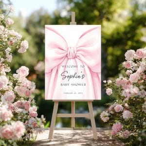 May include: A baby shower welcome sign on a wooden easel. The sign features a large pink bow design and the words "Welcome to Sophia's Baby Shower" in elegant script. The date is February 24, 20XX.