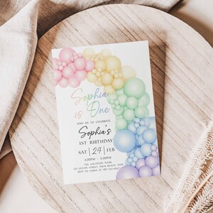 May include: A birthday invitation featuring a colorful balloon arch in pastel shades. The text "Sophia is One" is written in a script font, with additional details about the event. The invitation is set on a wooden surface.