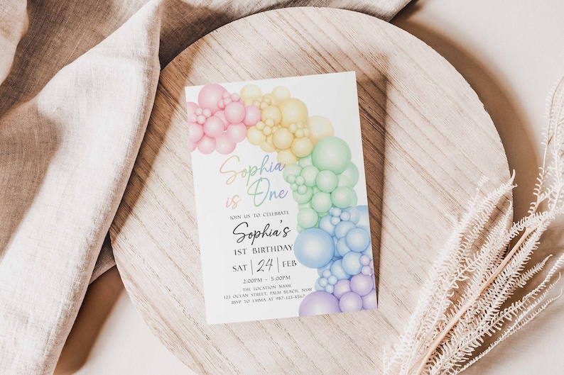 May include: A birthday invitation featuring a colorful balloon arch in pastel shades. The text "Sophia is One" is written in a script font, with additional details about the event. The invitation is set on a wooden surface.