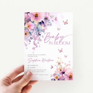 Może przedstawiać: Zaproszenie na baby shower z kwiatowym wzorem. Zaproszenie zawiera akwarelowe kwiaty w odcieniach r&oacute;żu, fioletu i ż&oacute;łci, z motylami. Tekst brzmi "Baby in Bloom" i zawiera szczeg&oacute;ły dotyczące wydarzenia.