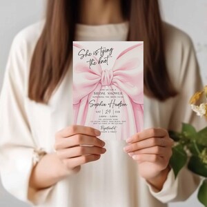 K&ouml;nnte beinhalten: Eine rosa Brautparty-Einladung mit einem Aquarell-Schleifendesign. Die Einladung enth&auml;lt den Text "She is tying the knot" und Details zur Veranstaltung, einschlie&szlig;lich des Namens "Sophie Hudson" und des Datums.