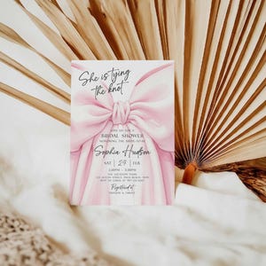 K&ouml;nnte beinhalten: Einladungskarte zur Brautparty mit einem gro&szlig;en rosa Schleifendesign. Der Text "She is tying the knot" steht oben, mit Details zur Veranstaltung, einschlie&szlig;lich Datum, Uhrzeit und Ort. Die Einladung befindet sich vor einem Hintergrund aus getrockneten Palmbl&auml;ttern.