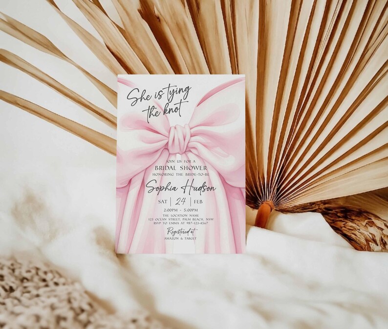 K&ouml;nnte beinhalten: Einladungskarte zur Brautparty mit einem gro&szlig;en rosa Schleifendesign. Der Text "She is tying the knot" steht oben, mit Details zur Veranstaltung, einschlie&szlig;lich Datum, Uhrzeit und Ort. Die Einladung befindet sich vor einem Hintergrund aus getrockneten Palmbl&auml;ttern.