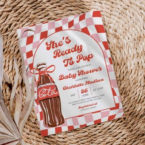 May include: Baby shower invitation with a retro design. The invitation features a red and pink checkered pattern and a vintage illustration of a cola bottle. Text includes "She's Ready To Pop" and event details.