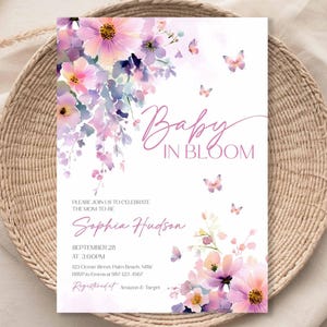Może przedstawiać: Zaproszenie na baby shower ze słowami "Baby in Bloom" w r&oacute;żowym skrypcie. Zaproszenie zawiera akwarelowe ilustracje kwiat&oacute;w i motyli w odcieniach r&oacute;żu i fioletu. Szczeg&oacute;ły obejmują imię przyszłej mamy, datę, godzinę i miejsce.