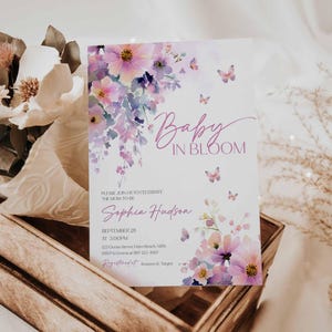 Może przedstawiać: Zaproszenie na baby shower z kwiatowym wzorem akwarelowym. Zaproszenie zawiera tekst "Baby in Bloom" i imię "Sophia Hudson". Zaproszenie jest ozdobione r&oacute;żowymi i fioletowymi kwiatami i motylami. Zaproszenie jest wyświetlane w drewnianej skrzyni.