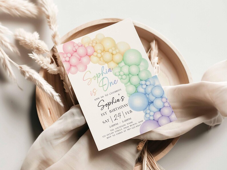 May include: A pastel-colored invitation card for a first birthday celebration. The card features a rainbow-colored bubble design and the text "Sophia is One". The event details are also included on the card.