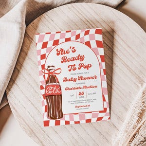 May include: A baby shower invitation with a retro design. The invitation features the text "She's Ready To Pop" and a cartoon illustration of a cola bottle. The background has a red and pink checkered pattern. The invitation is on a wooden surface.