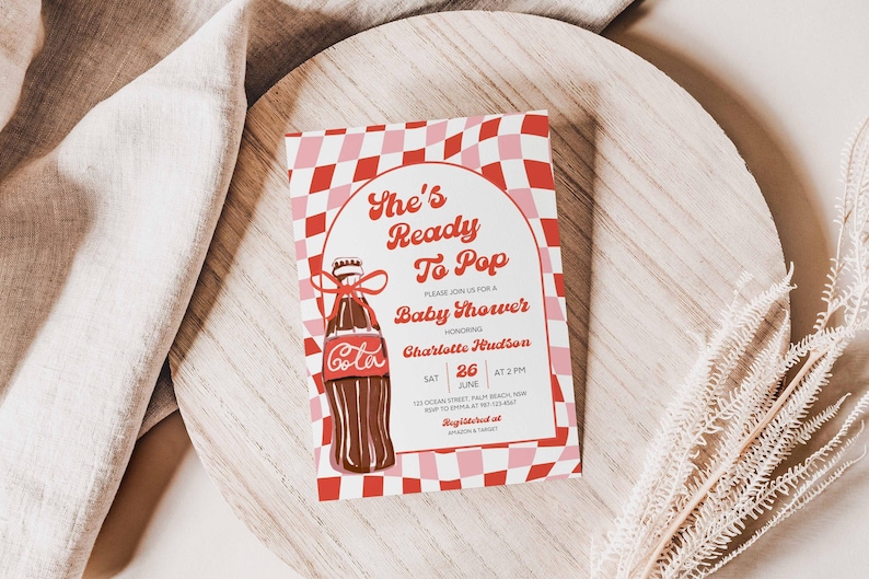 May include: A baby shower invitation with a retro design. The invitation features the text "She's Ready To Pop" and a cartoon illustration of a cola bottle. The background has a red and pink checkered pattern. The invitation is on a wooden surface.