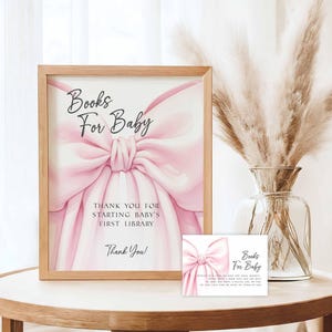 May include: A framed sign and a card with the text "Books For Baby". The sign features a pink bow illustration and the text "THANK YOU FOR STARTING BABY'S FIRST LIBRARY". The card has the same bow design and text. The items are on a wooden table.