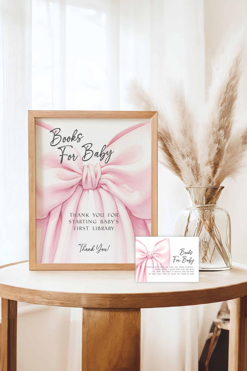 May include: A framed sign and a card with the text "Books For Baby". The sign features a pink bow illustration and the text "THANK YOU FOR STARTING BABY'S FIRST LIBRARY". The card has the same bow design and text. The items are on a wooden table.